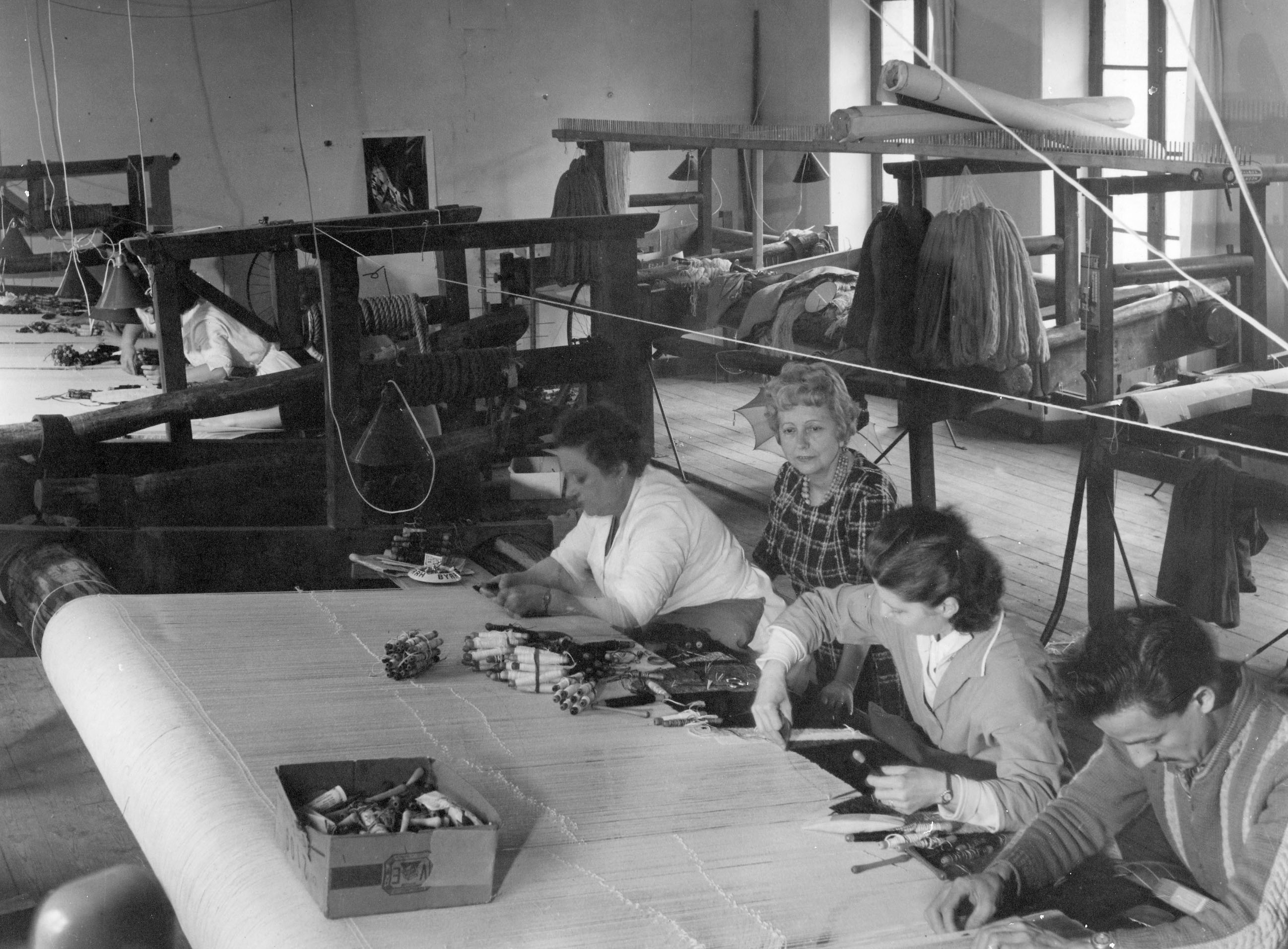 Vue de l'atelier du 1er étage de la manufacture Goubely, avec les ouvriers au travail et Suzanne, en arrière-plan, surveillant la progression du tissage sur des métiers à barre (v. 1970)