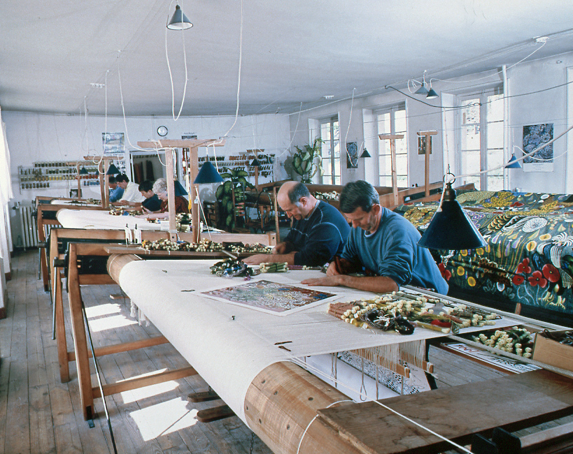 Ouvriers au travail, dans l'atelier de tissage du premier étage de la manufacture. A l'arrière-plan, une tapisserie achevée d'après un carton de Dom Robert (v. 1970)