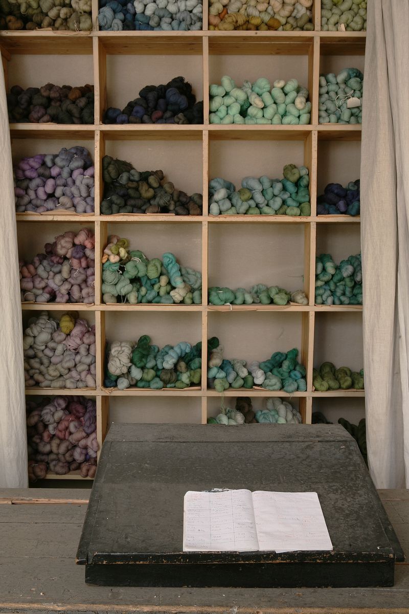 Détail des casiers contenant les écheveaux de laine vierge, rangés par nuances de couleur, dans le magasin des laines, au second étage, avec un registre de comptabilité de l'entreprise, ouvert, au premier plan.