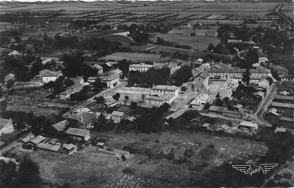 Carte postale (collection particulière) : vue aérienne du village, baraquements construits sur la place, rue du 19 mars 1962 (après guerre, le clocher de l'église est en ruine).