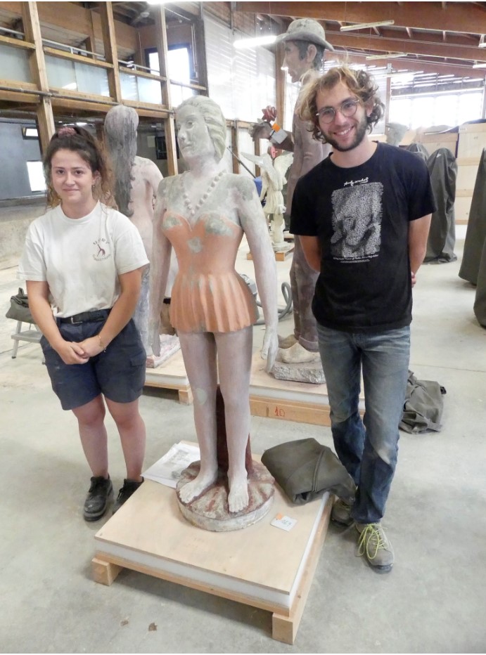 Sculpture de femme à la robe orange après la première semaine de chantier, encadrée par les deux étudiants chargés de sa restauration.